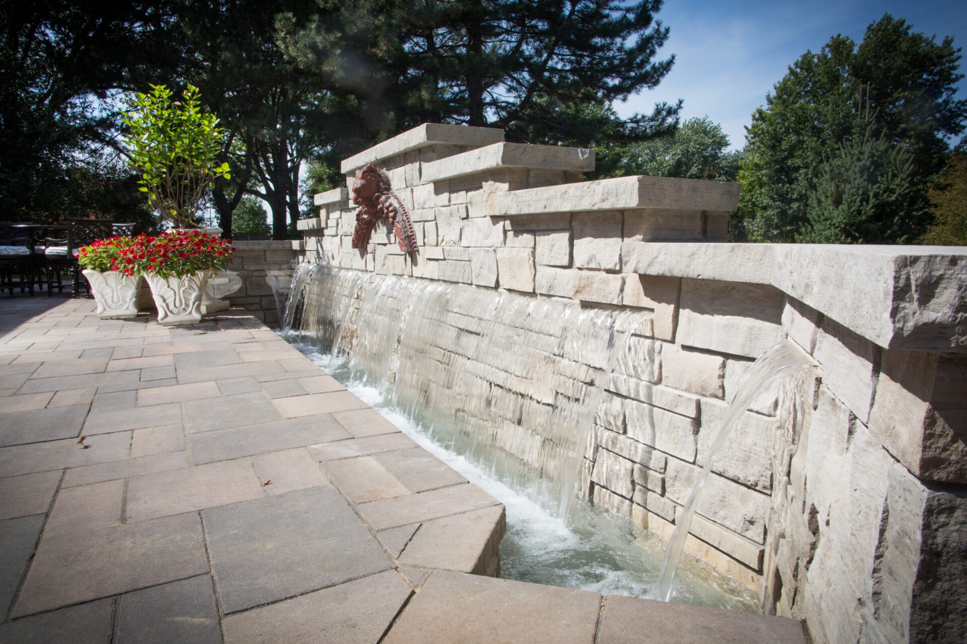 Stone fountain with cascading water, surrounded by trees and floral planters, on a tiled patio. Sunny day with clear skies.