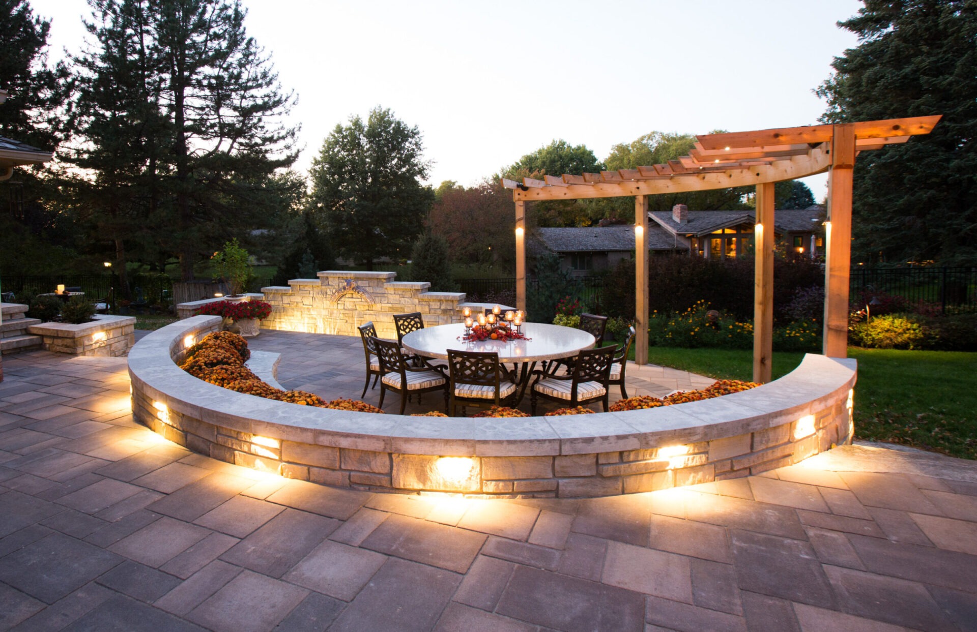 Elegant backyard patio with circular dining area, warm lighting, stone walls, pergola, and lush landscaping, creating a cozy outdoor ambiance at dusk.