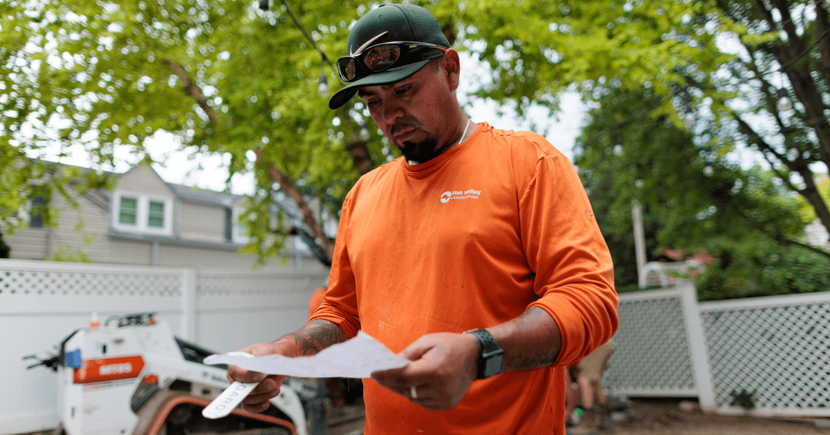 A person wearing an orange shirt examines a paper outdoors, near construction equipment and a white fence, surrounded by green trees.