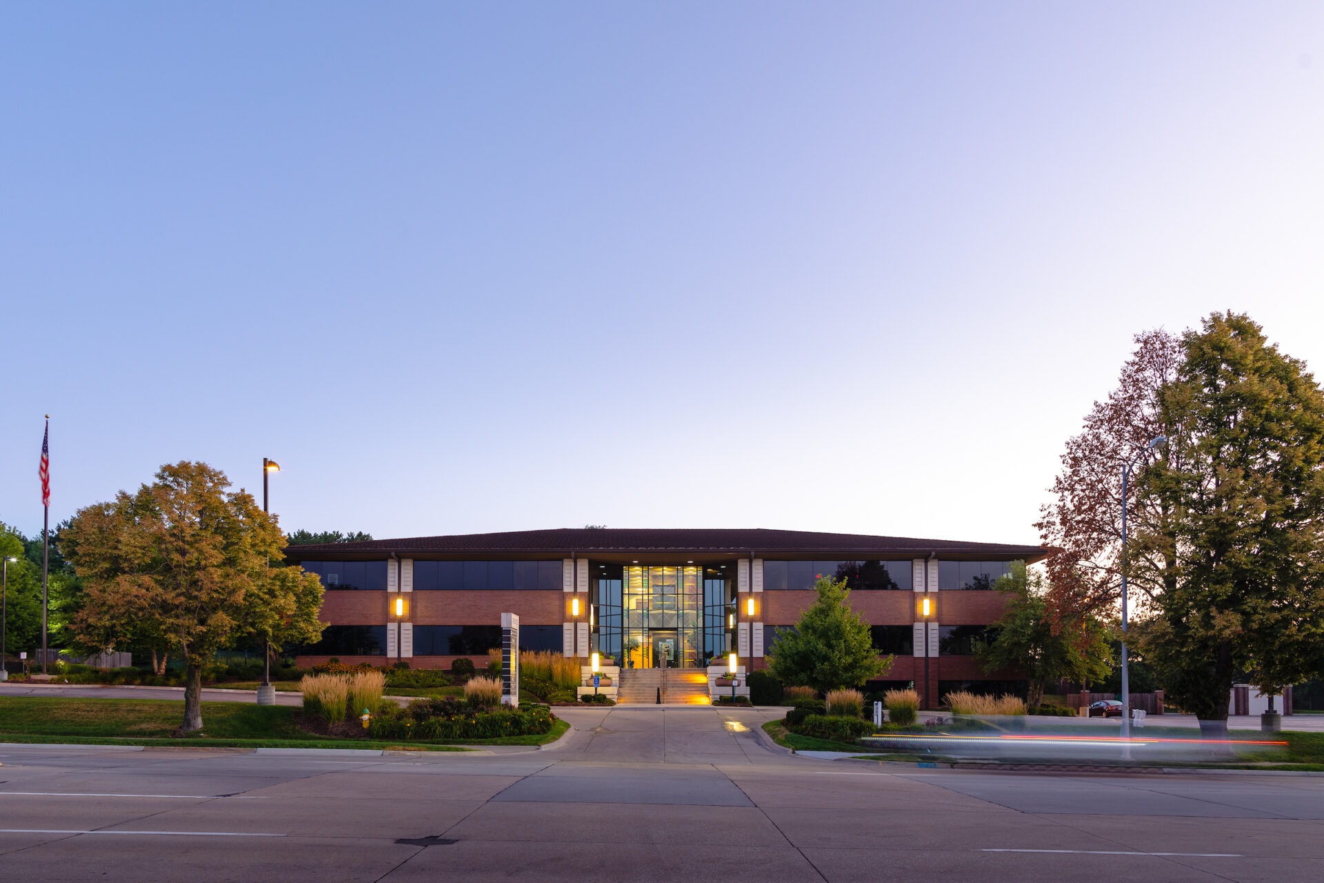 Modern office building at dusk with illuminated windows, surrounded by trees and landscaped gardens, an American flag visible on the left.