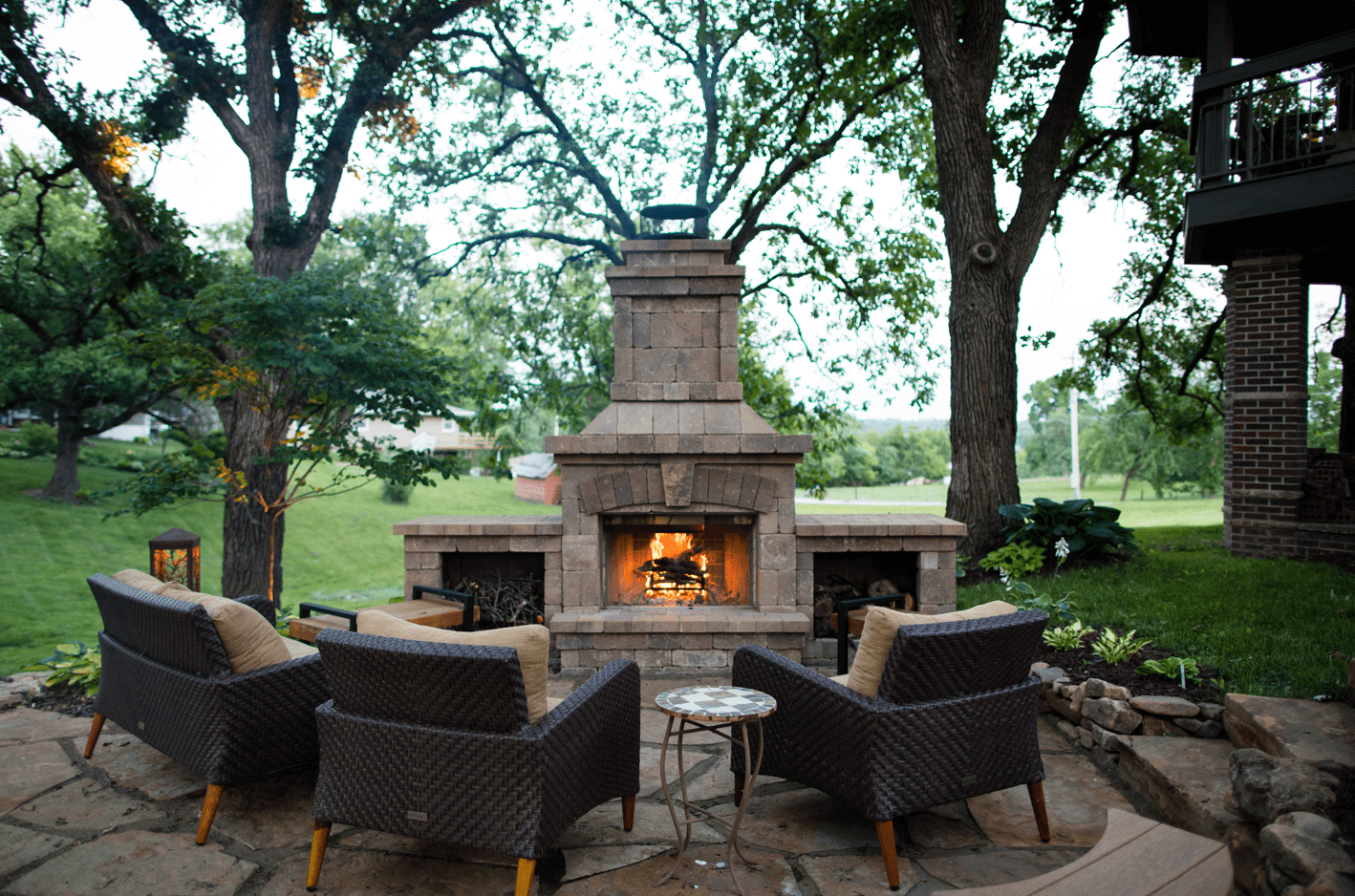 Cozy outdoor patio with a brick fireplace, surrounded by trees and wicker chairs, creating a serene evening ambiance in lush green garden.