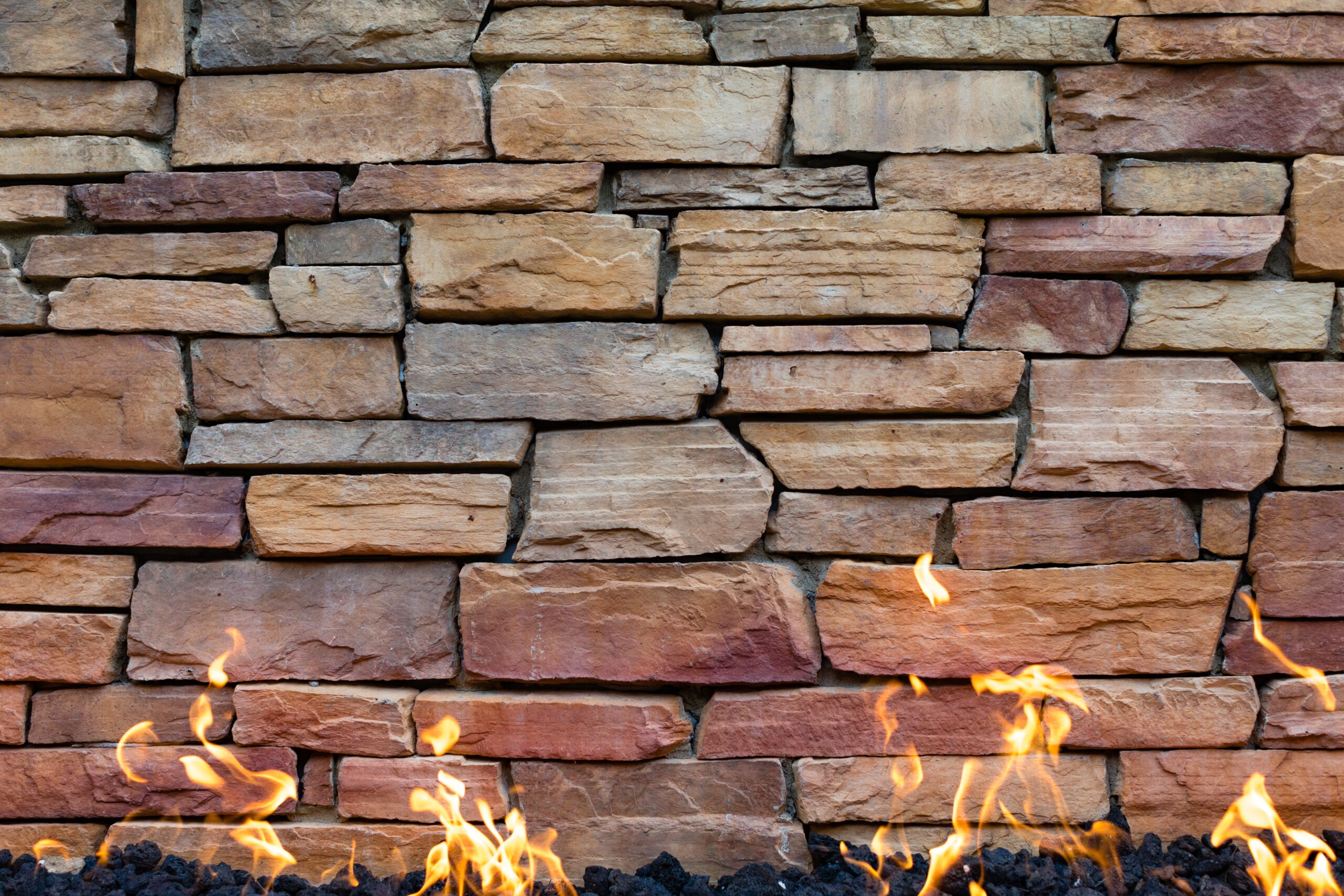 A rustic stone wall with orange, red, and brown hues has vivid flames flickering at the base, creating a warm, cozy ambiance.