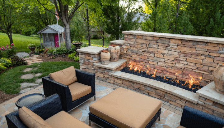 A cozy outdoor patio with wicker furniture, a stone fireplace, lush greenery, and a small wooden playhouse nestled among trees in the background.