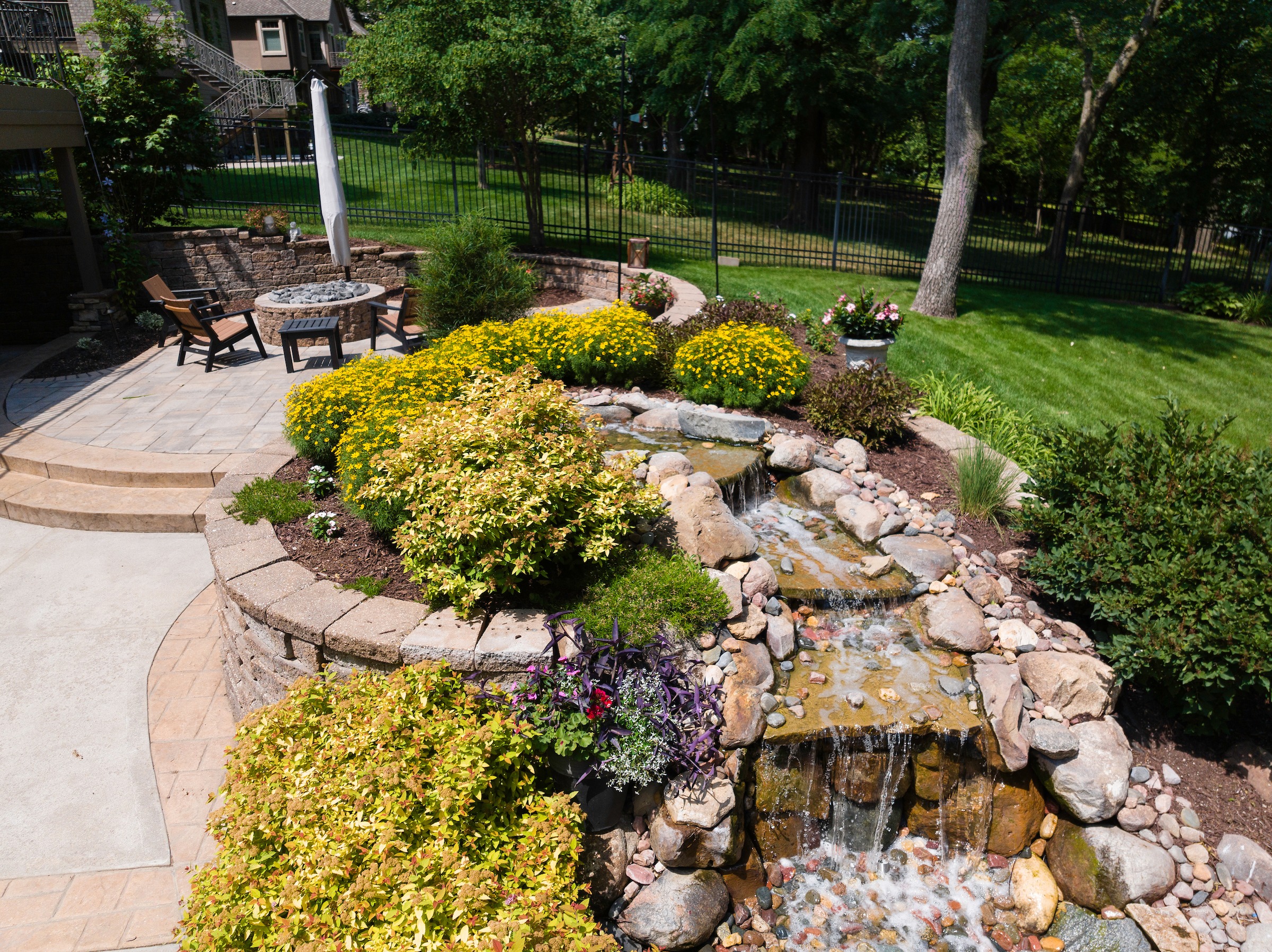 A landscaped backyard features a small waterfall, stone patio, seating area with a table, vibrant flowers, and lush greenery under bright sunshine.