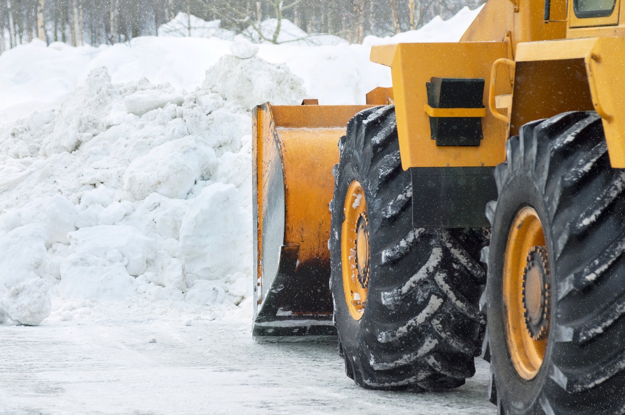 A yellow snow plow with large tires is clearing a path through a snowy landscape, surrounded by piled snow.