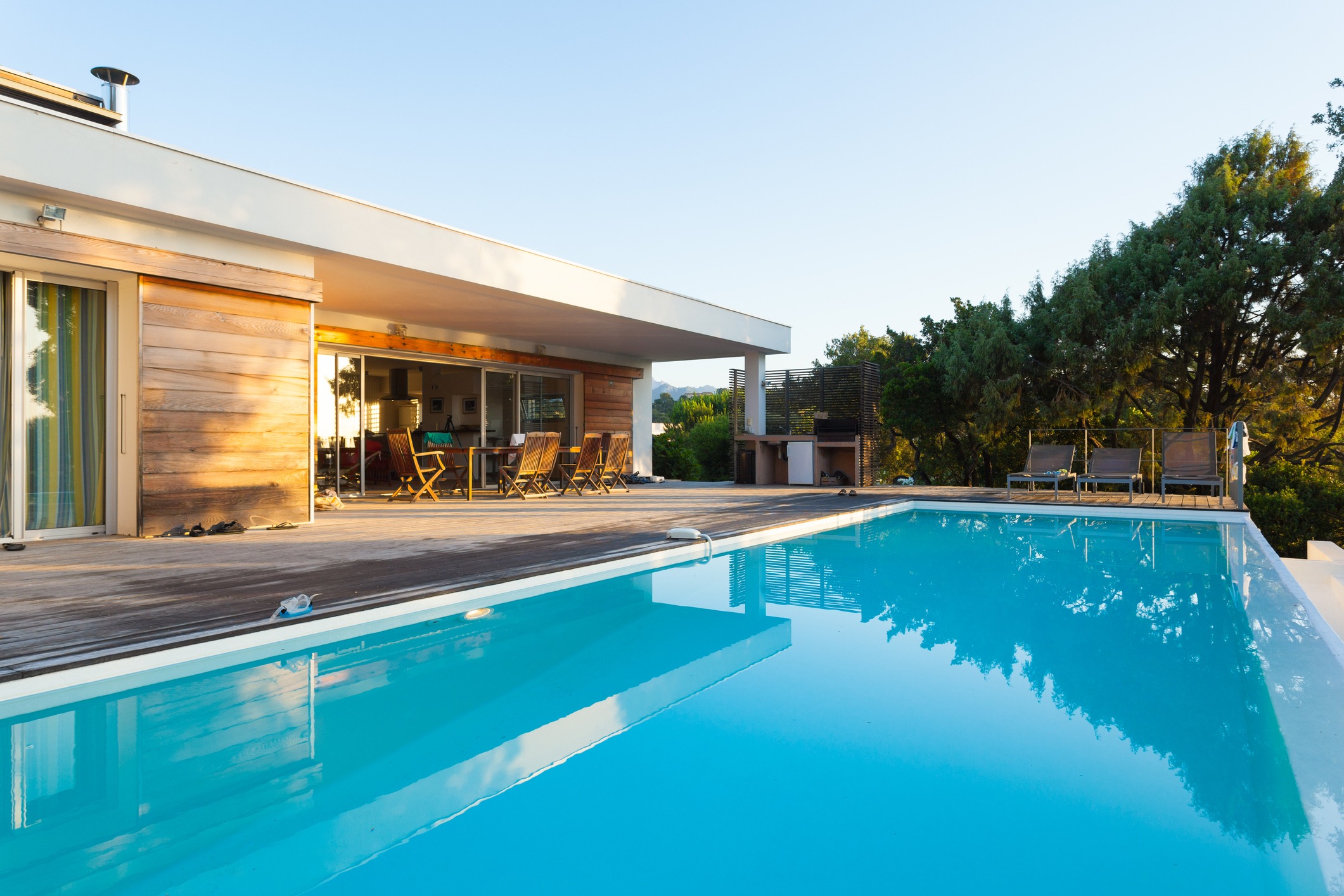 Modern house with a wooden deck overlooks a clear swimming pool. Outdoor seating and lush greenery surround the area, creating a serene atmosphere.