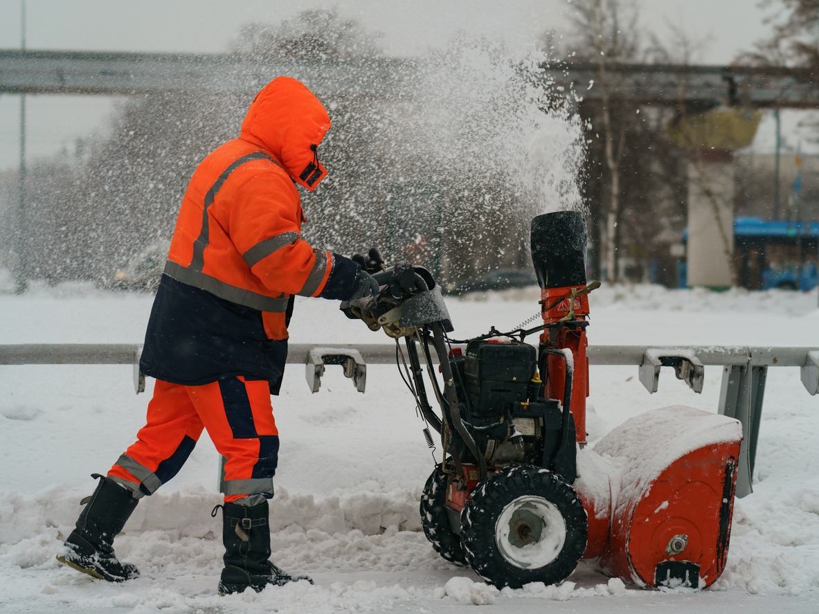 A person wearing bright orange winter gear operates a snowblower, clearing a path through heavy snowfall near a snowy road or path.