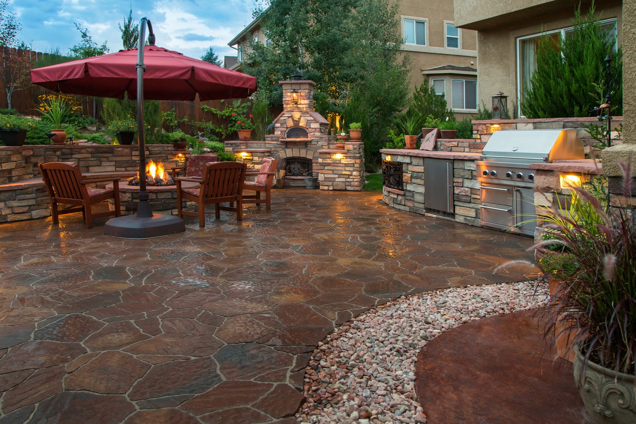 A cozy backyard patio with a fire pit, stone fireplace, outdoor kitchen, and colorful umbrella, surrounded by lush garden greenery.