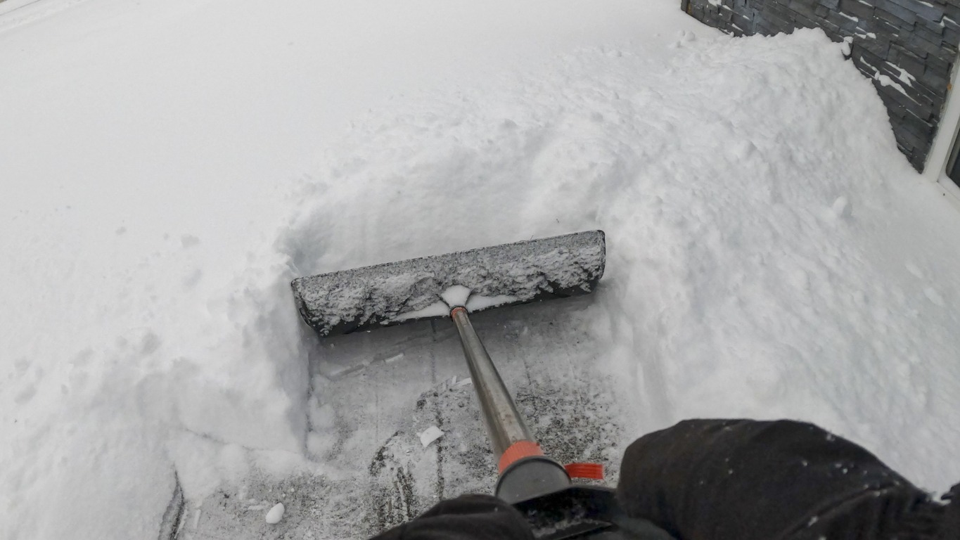 A person uses a snow shovel to clear a path surrounded by deep snow near a brick wall on a cold, snowy day.