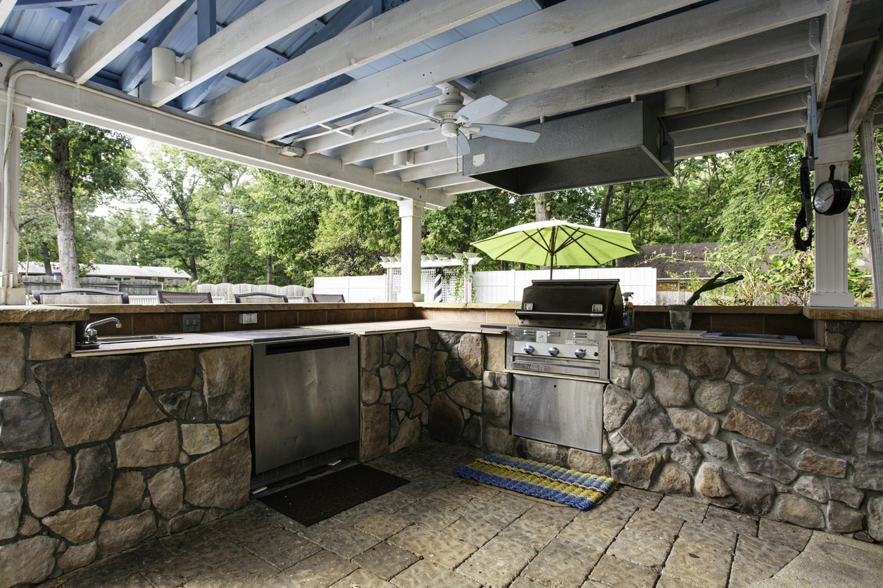 Outdoor kitchen with stone countertops, grill, and green umbrella, under a wooden pergola. Surrounded by trees and fencing for privacy.