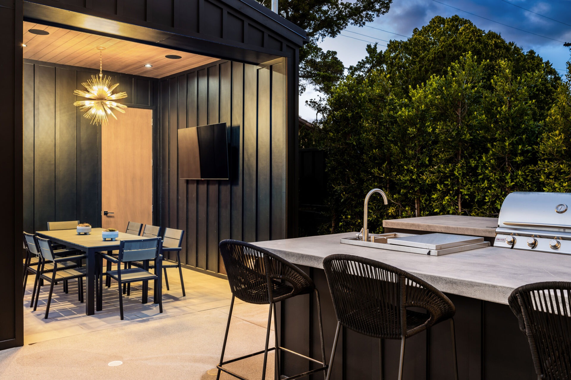 Modern outdoor patio featuring a dining area with a contemporary light fixture, barbecue grill, and bar seating, surrounded by lush greenery.