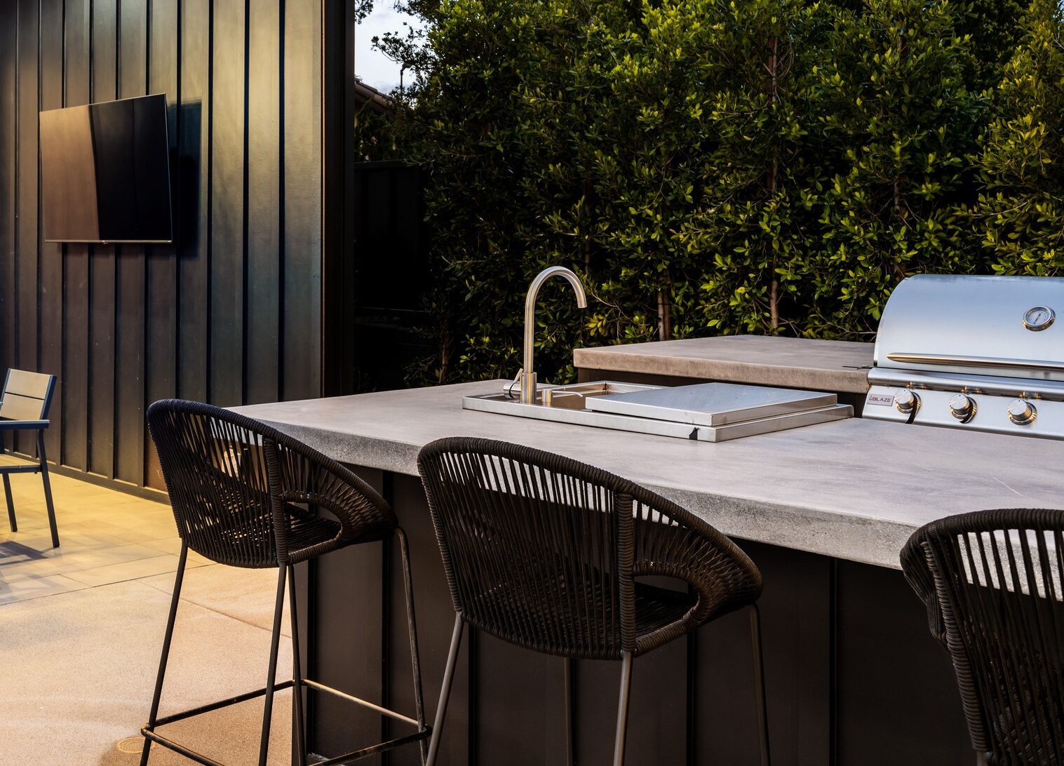 Modern outdoor patio featuring a dining area with a contemporary light fixture, barbecue grill, and bar seating, surrounded by lush greenery.