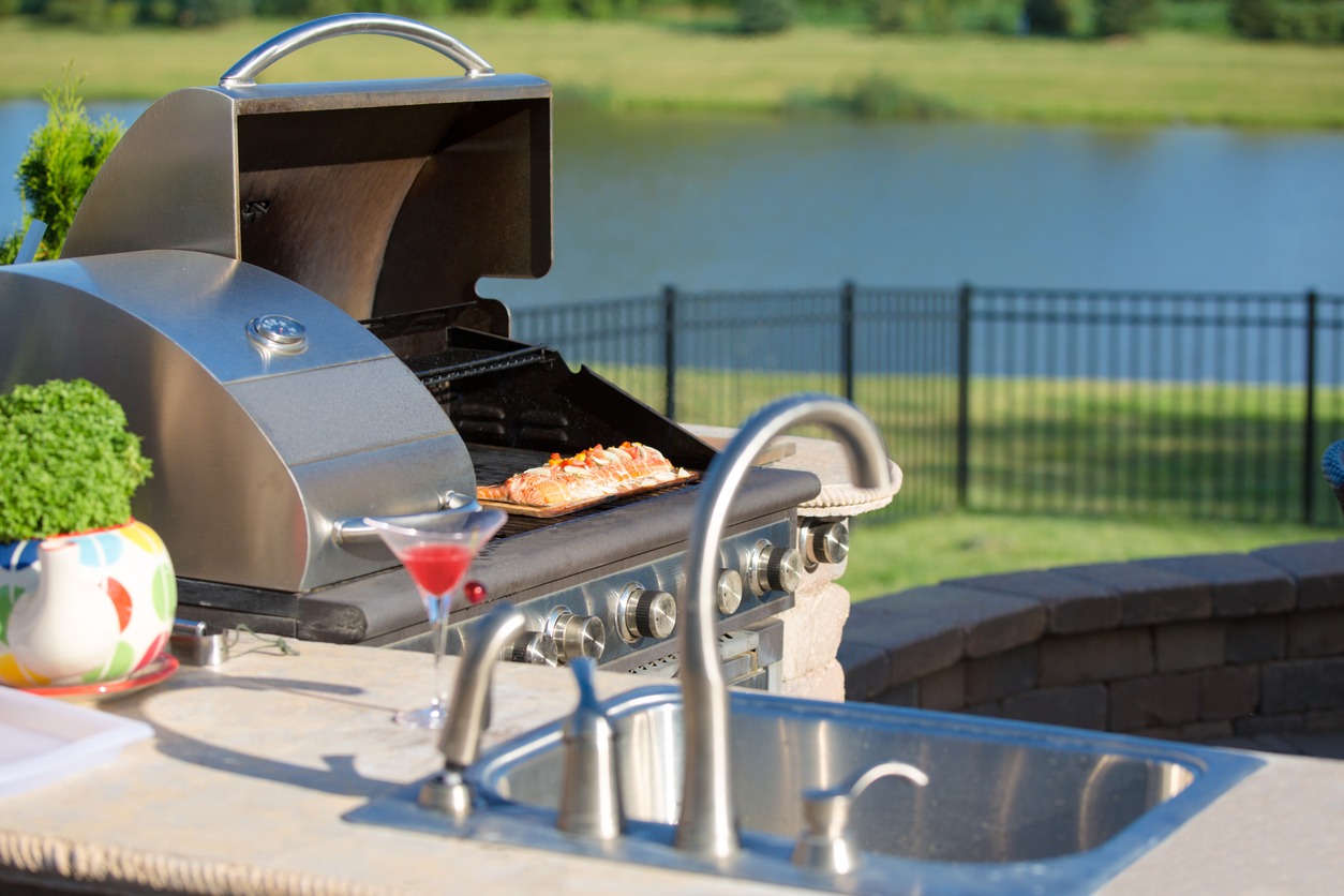 A barbecue grill with salmon, cocktail, and colorful vase beside a sink, overlooking a fenced lawn and lake in the background.