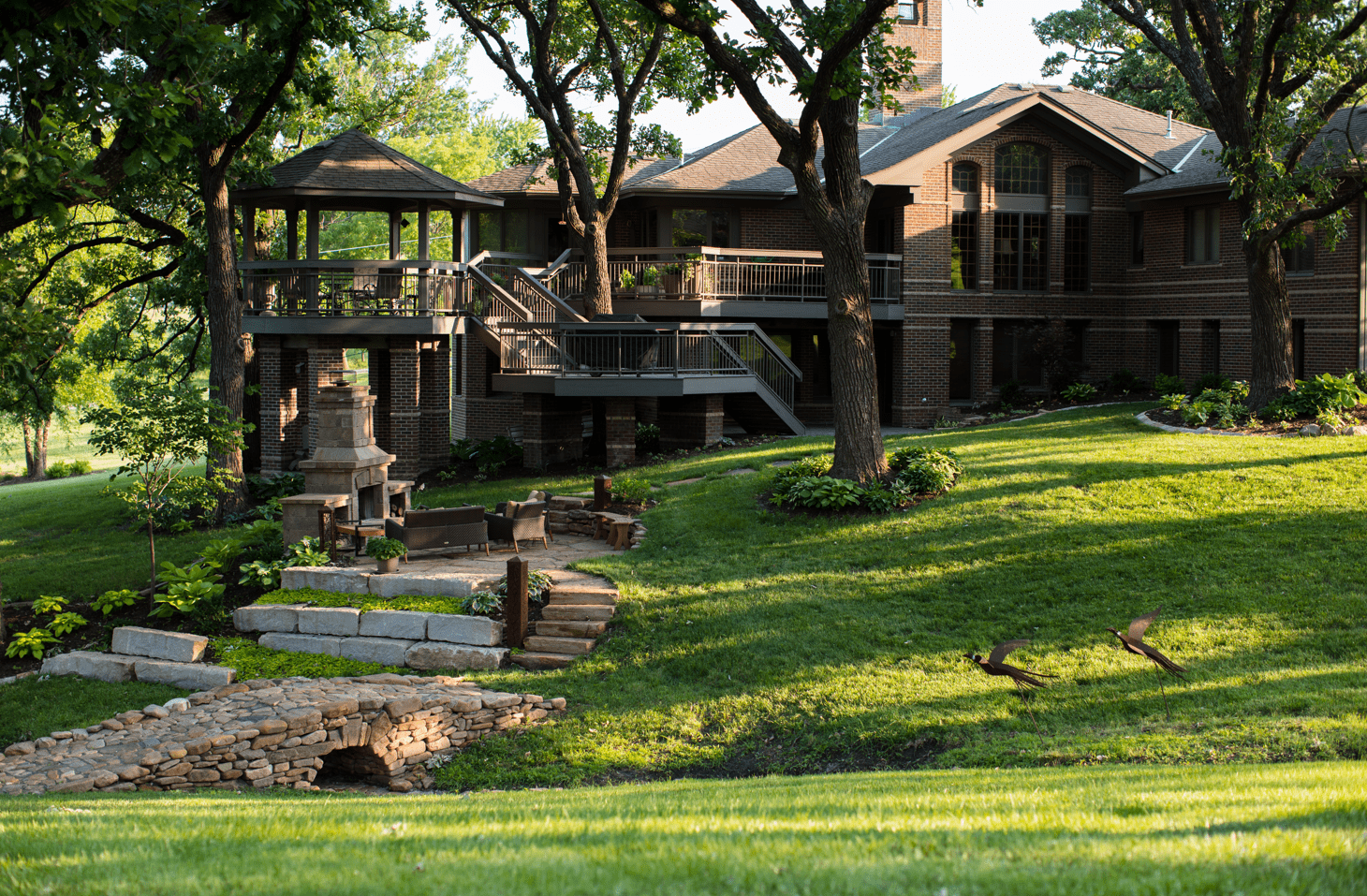 A brick house with wraparound balcony, surrounded by trees and well-manicured lawn, features a stone bridge and outdoor seating area.