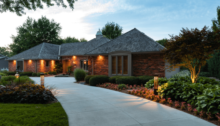 Cozy brick house with warm exterior lighting and landscaped garden lining a curved driveway, surrounded by lush greenery and evening skies.