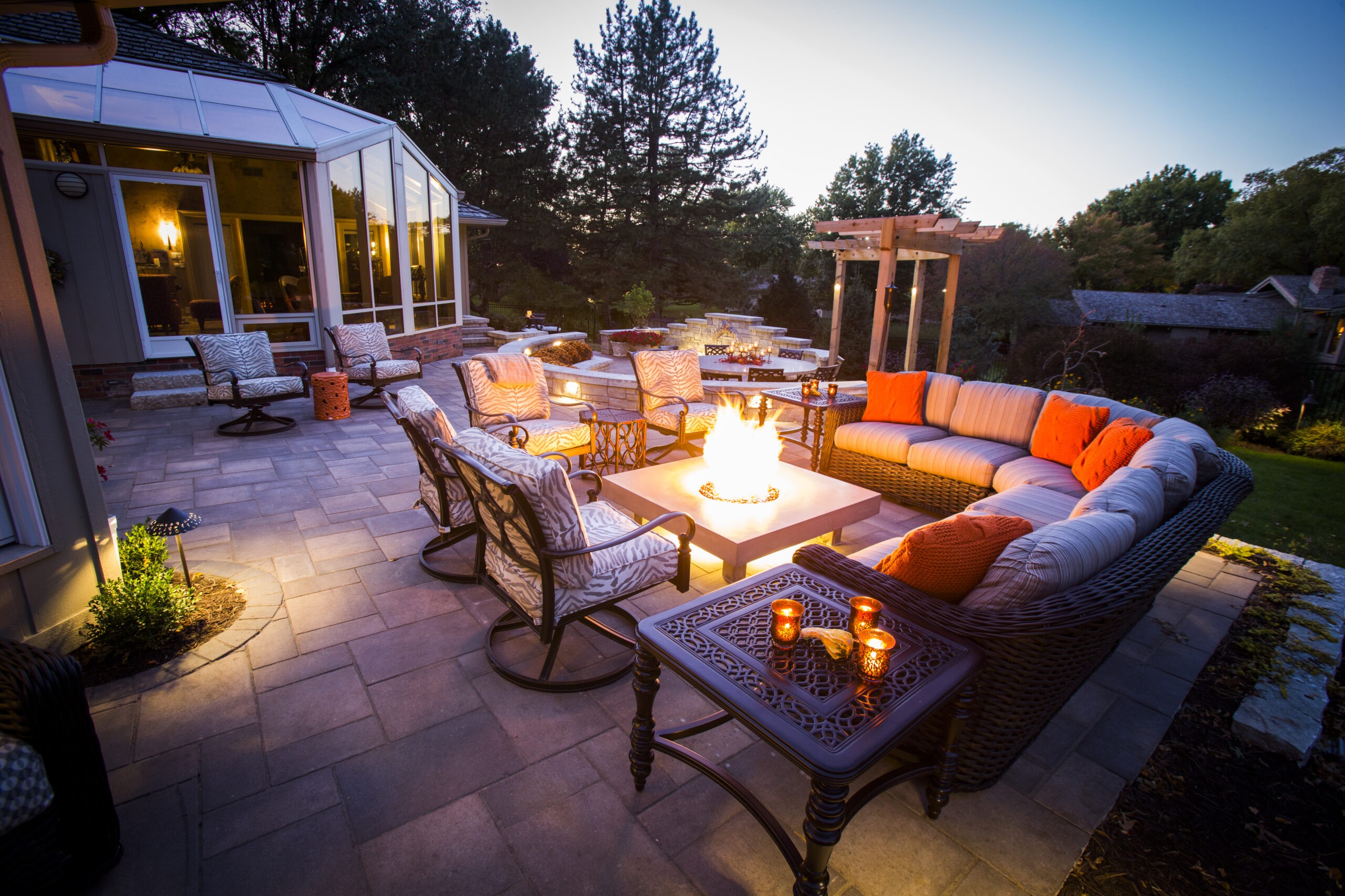 Cozy outdoor patio with elegant furniture, warm lighting, and a central firepit. Surrounded by lush trees, creating a serene evening atmosphere.