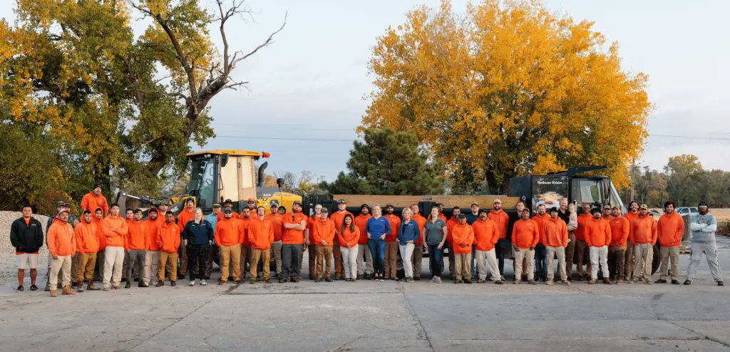 A group of people wearing orange jackets poses outdoors with construction equipment, surrounded by autumn trees on a clear day.