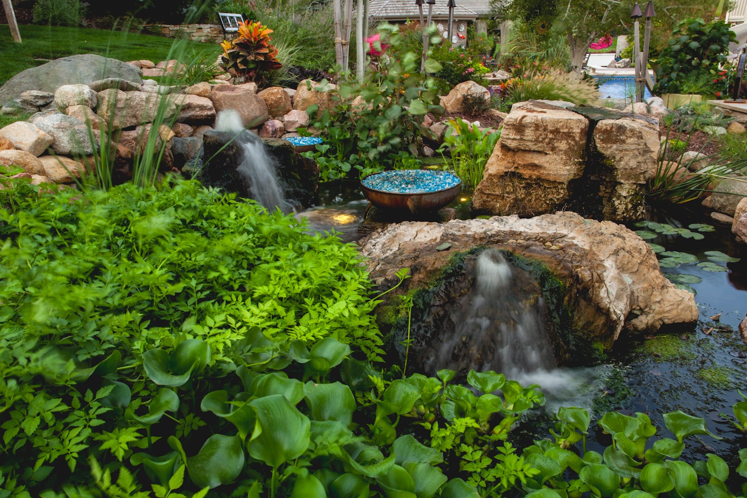 Natural-style pond with boulders, green plants, and dual waterfalls, enhanced with decorative bowls of stones.