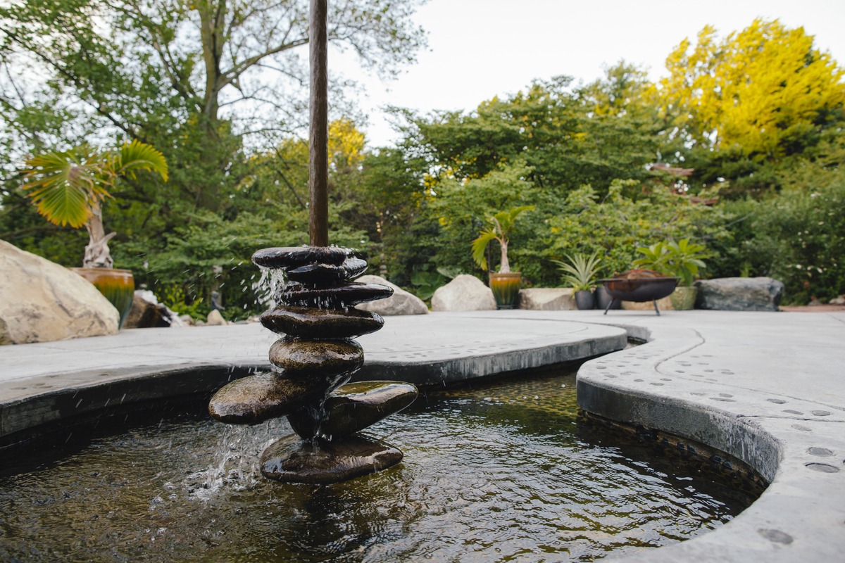 Artistic vertical fountain with flat stones stacked and water flowing into a small round pool, surrounded by lush greenery.