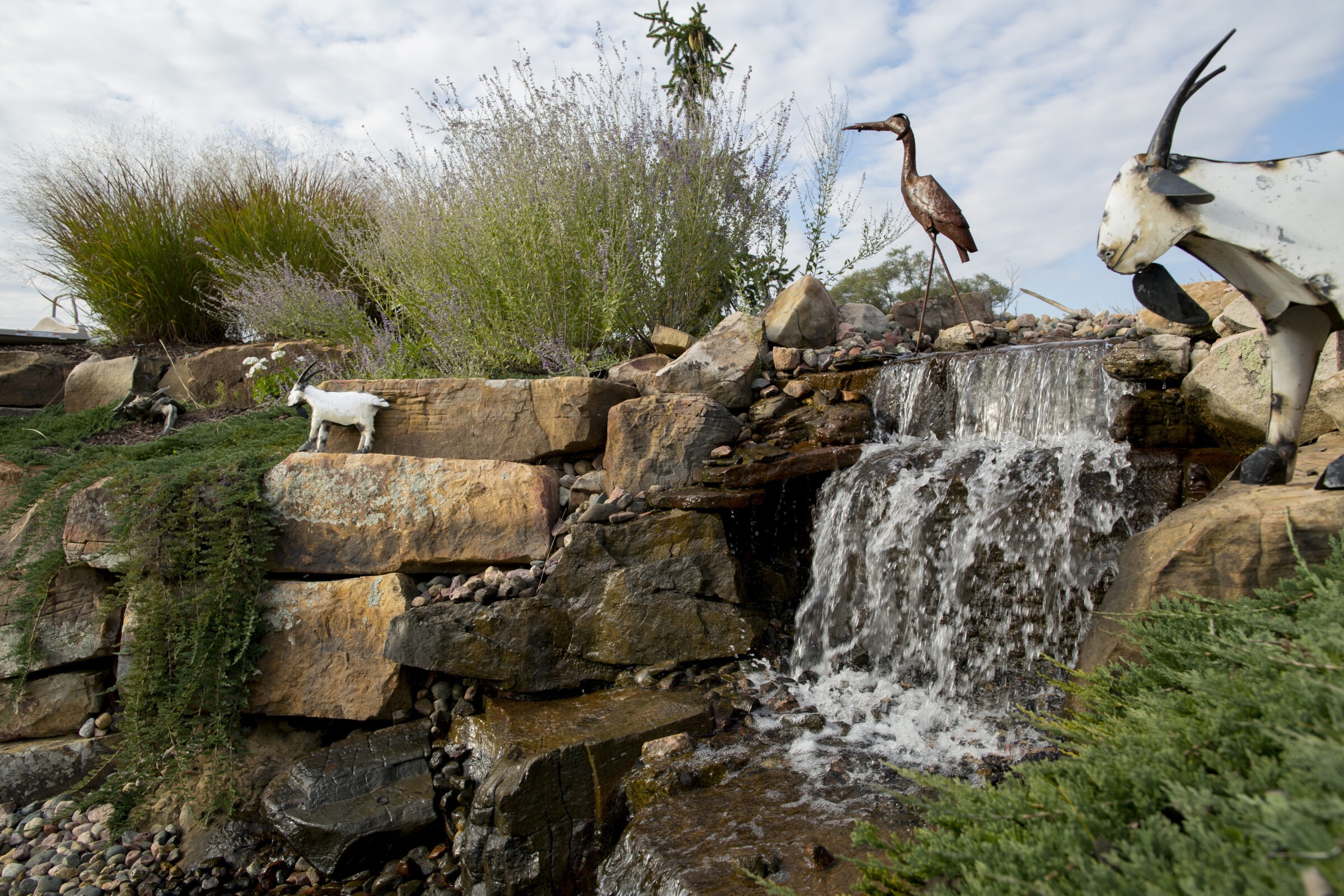 Garden waterfall cascading over natural stone with metal goat and heron sculptures as decorative accents.