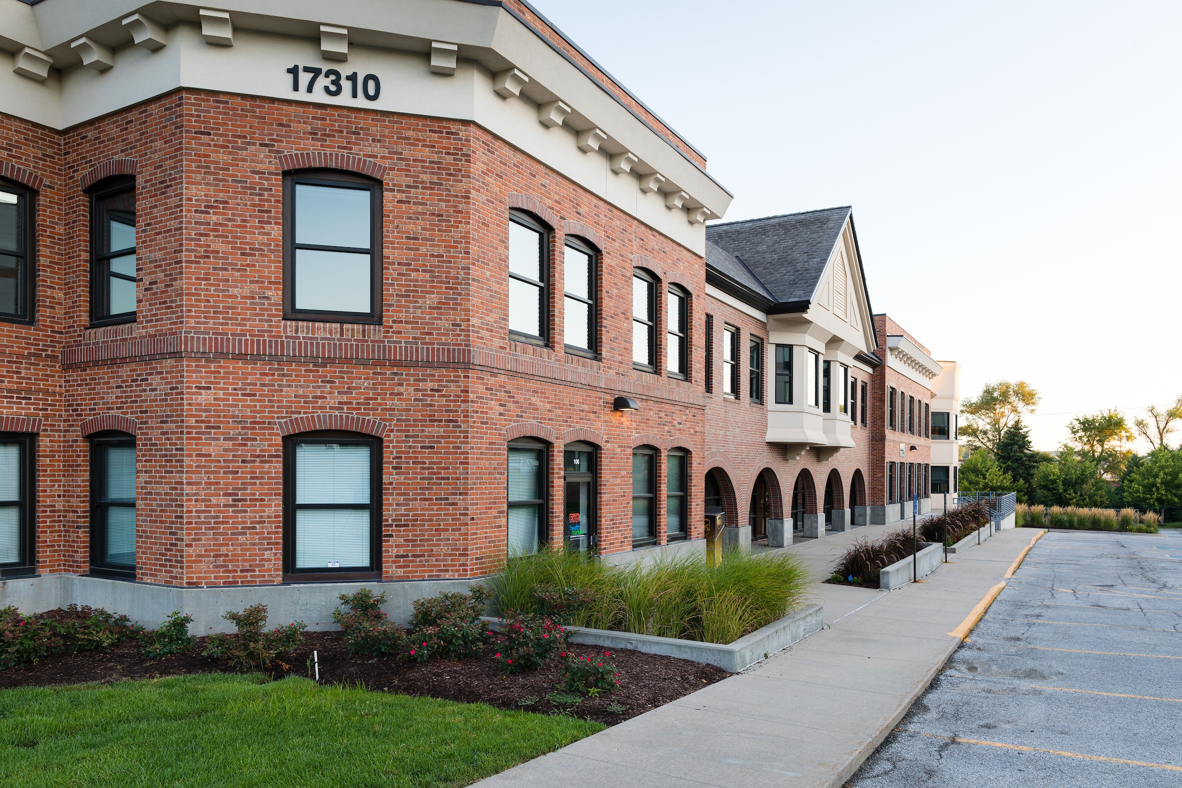 Red brick office building with arched windows, landscaped flower beds, and manicured lawn along the sidewalk.