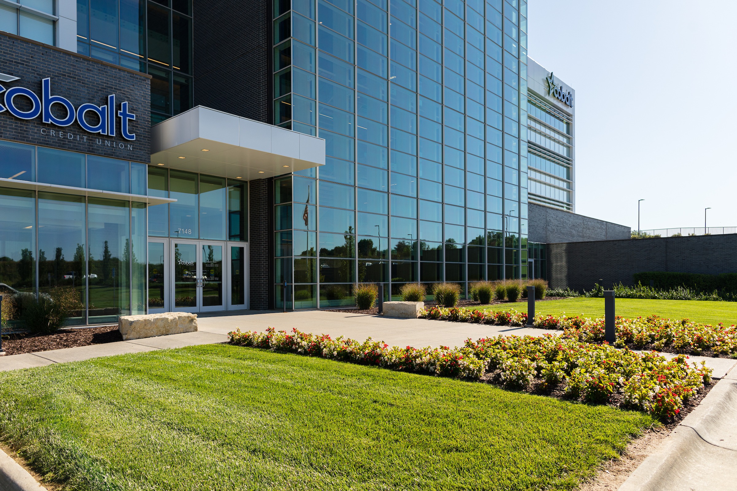 Exterior of a commercial building with tall glass façade, manicured lawn, and flower beds lining the entrance walkway