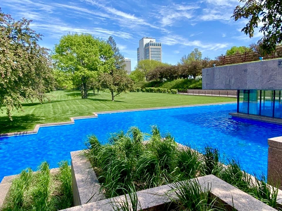Reflective blue pool with lush green lawns and city skyscraper visible in the distance.