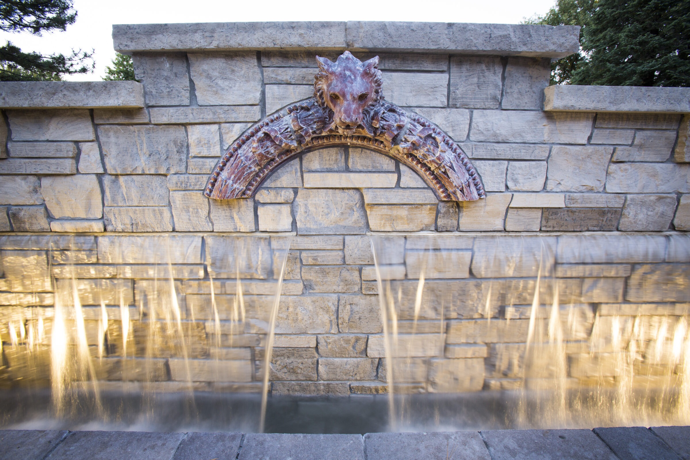 Decorative stone wall fountain with carved lion head detail and multiple streams of water illuminated by soft lighting, creating a dramatic effect.
