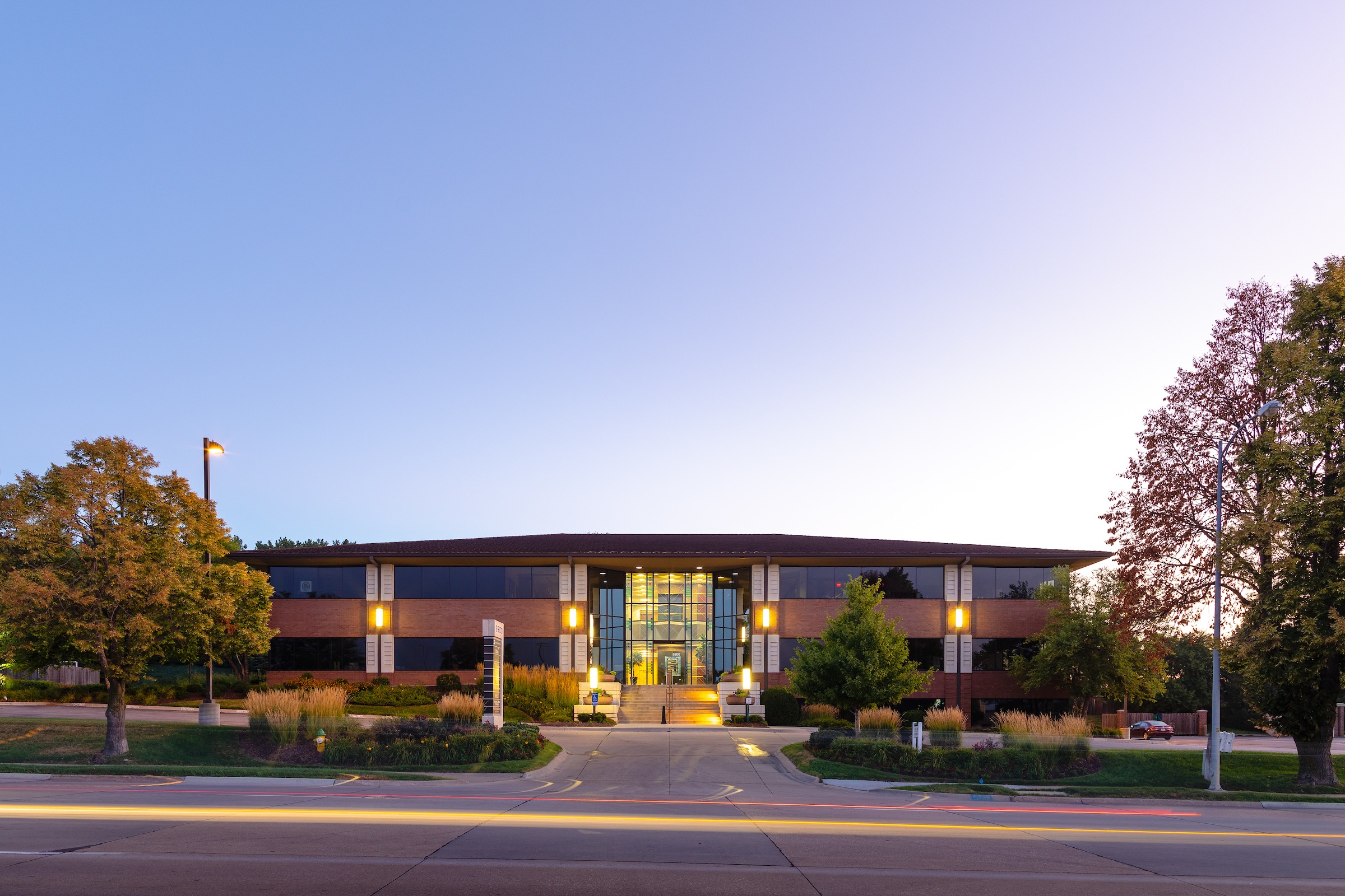 A symmetrical, two-story brick office building at dusk with a glass central entrance glowing warmly, flanked by trees and landscaped grounds, viewed from across a quiet street under a clear evening sky.