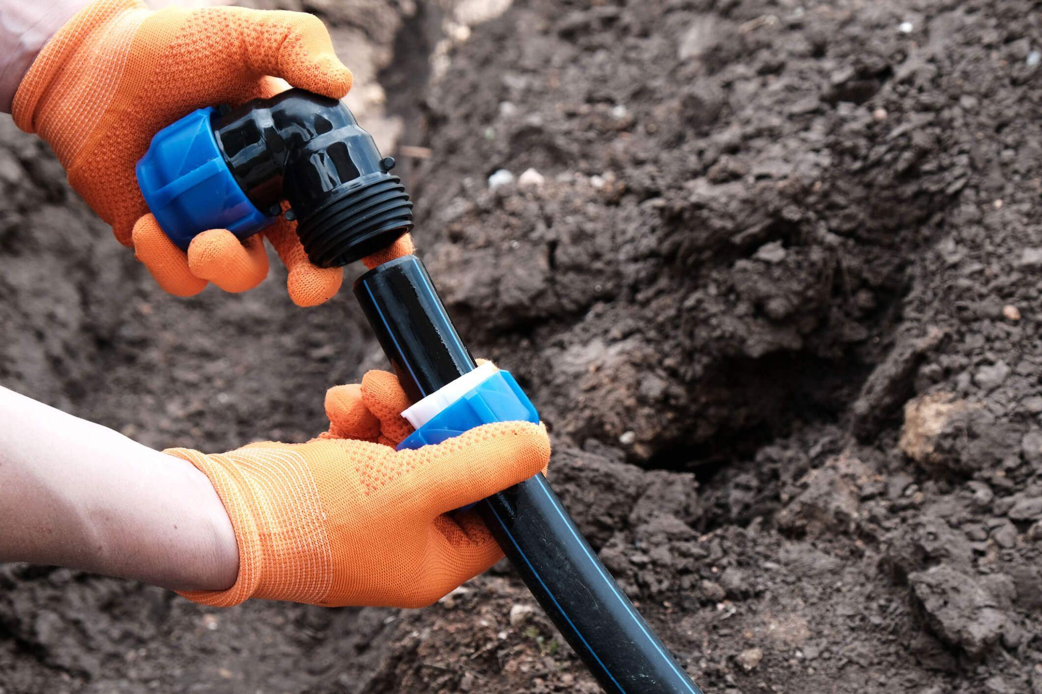 Hands wearing orange gloves connecting a black plastic irrigation pipe fitting in soil.