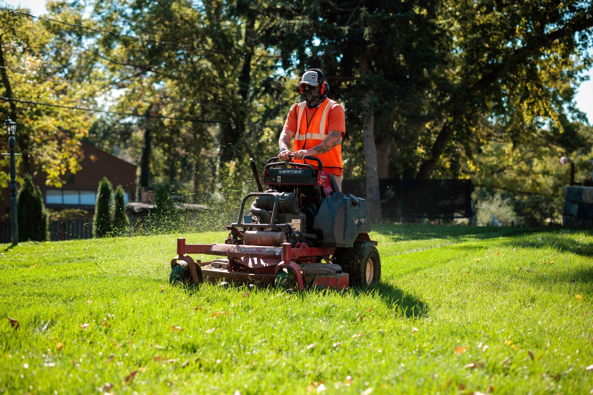 Worker in safety gear mowing a grassy area using a stand-on lawn mower.
