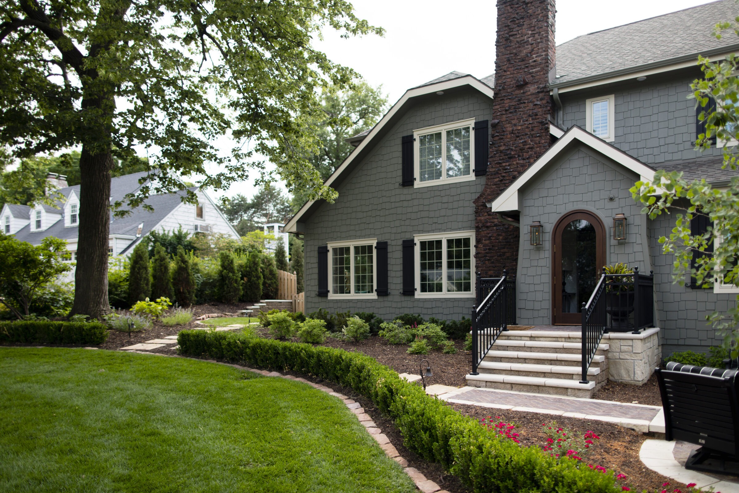 An exterior view of a two-story house with dark grey shingle siding, black shutters, and a tall brick chimney. A curved stone walkway leads up to a stone front porch with a dark arched door. The manicured front yard features a lush green lawn bordered by a neat, low boxwood hedge and fresh mulch beds filled with small shrubs.