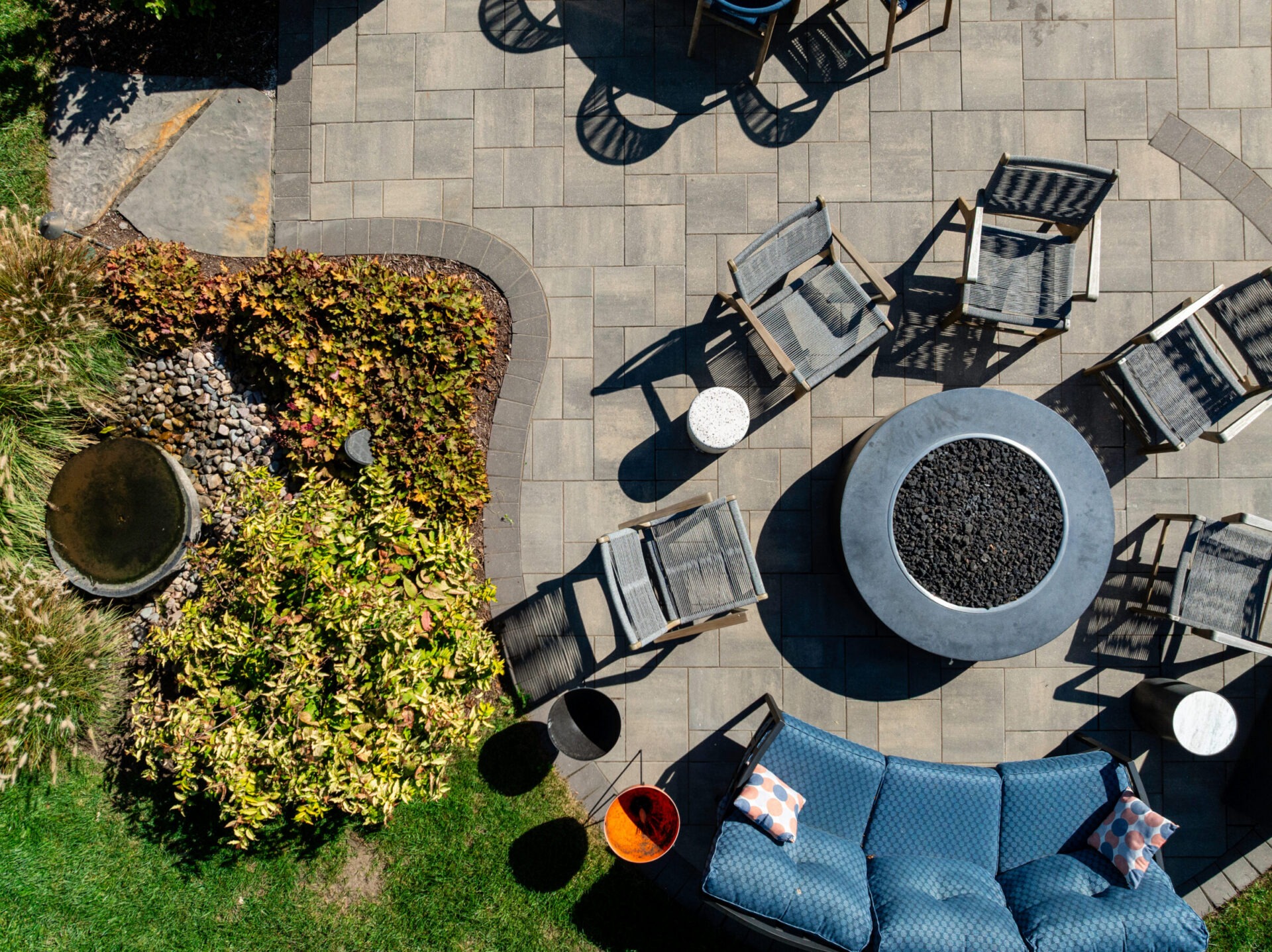 A high-angle, top-down view of a modern backyard patio paved with grey stone tiles. The space is arranged for entertaining, featuring a large, round grey stone fire pit surrounded by a blue curved outdoor sofa, several grey woven armchairs, and small accent tables. A landscaped garden bed with lush green and reddish foliage borders the patio.